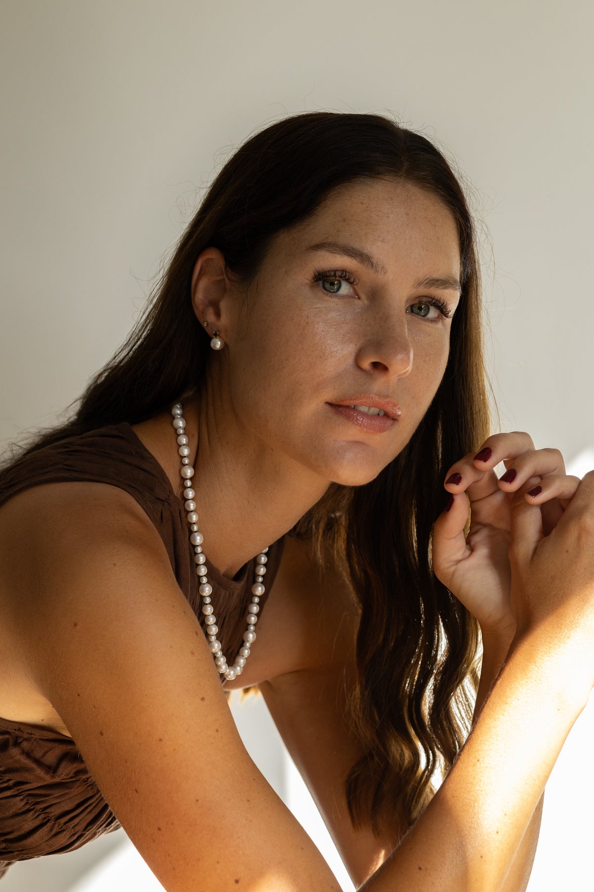 Woman with long dark hair and a pearl necklace sitting at a table with a warm light source.