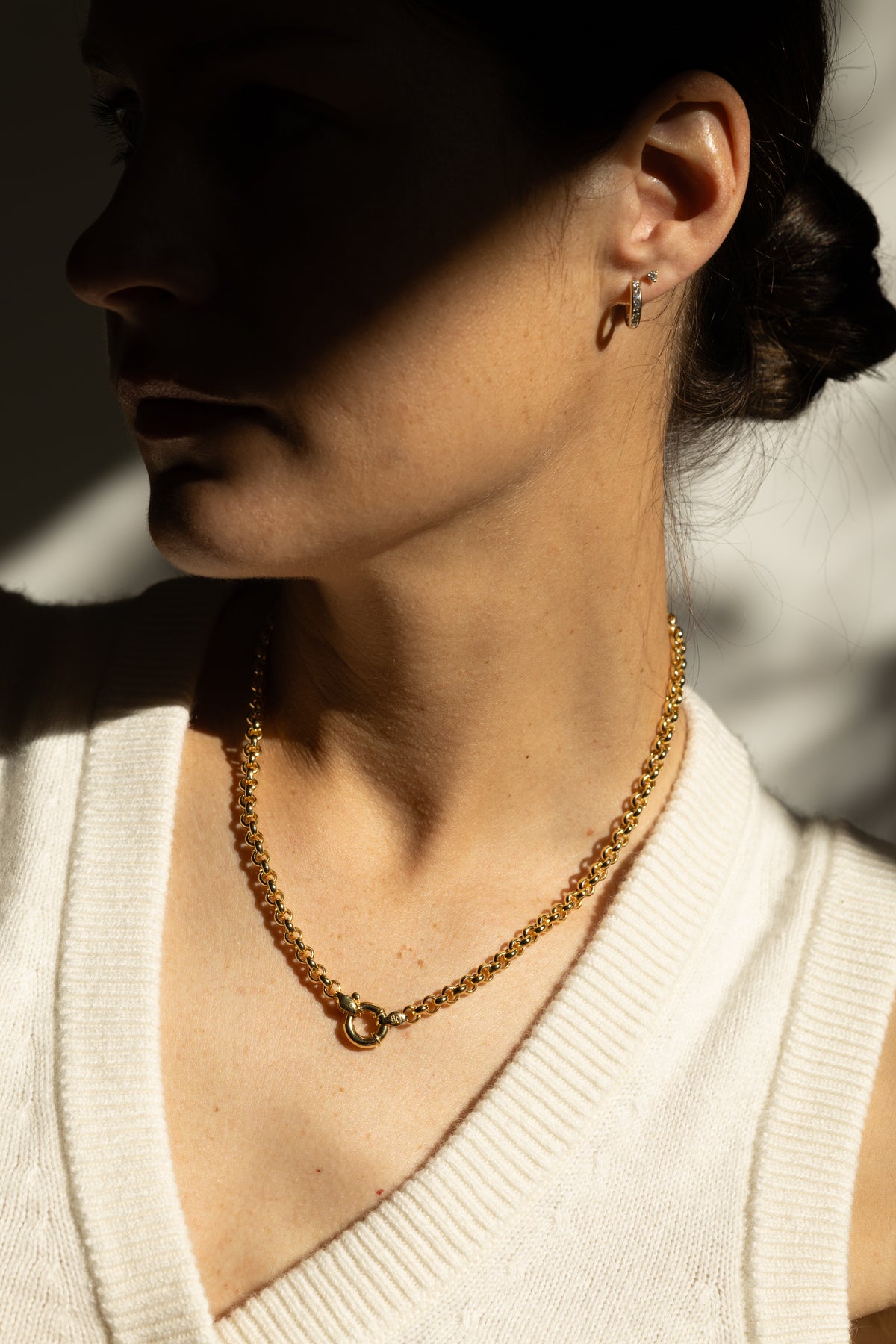 A close-up side profile of a woman wearing a statement gold chain necklace and diamond huggies, set against a high-contrast shadowed background.
