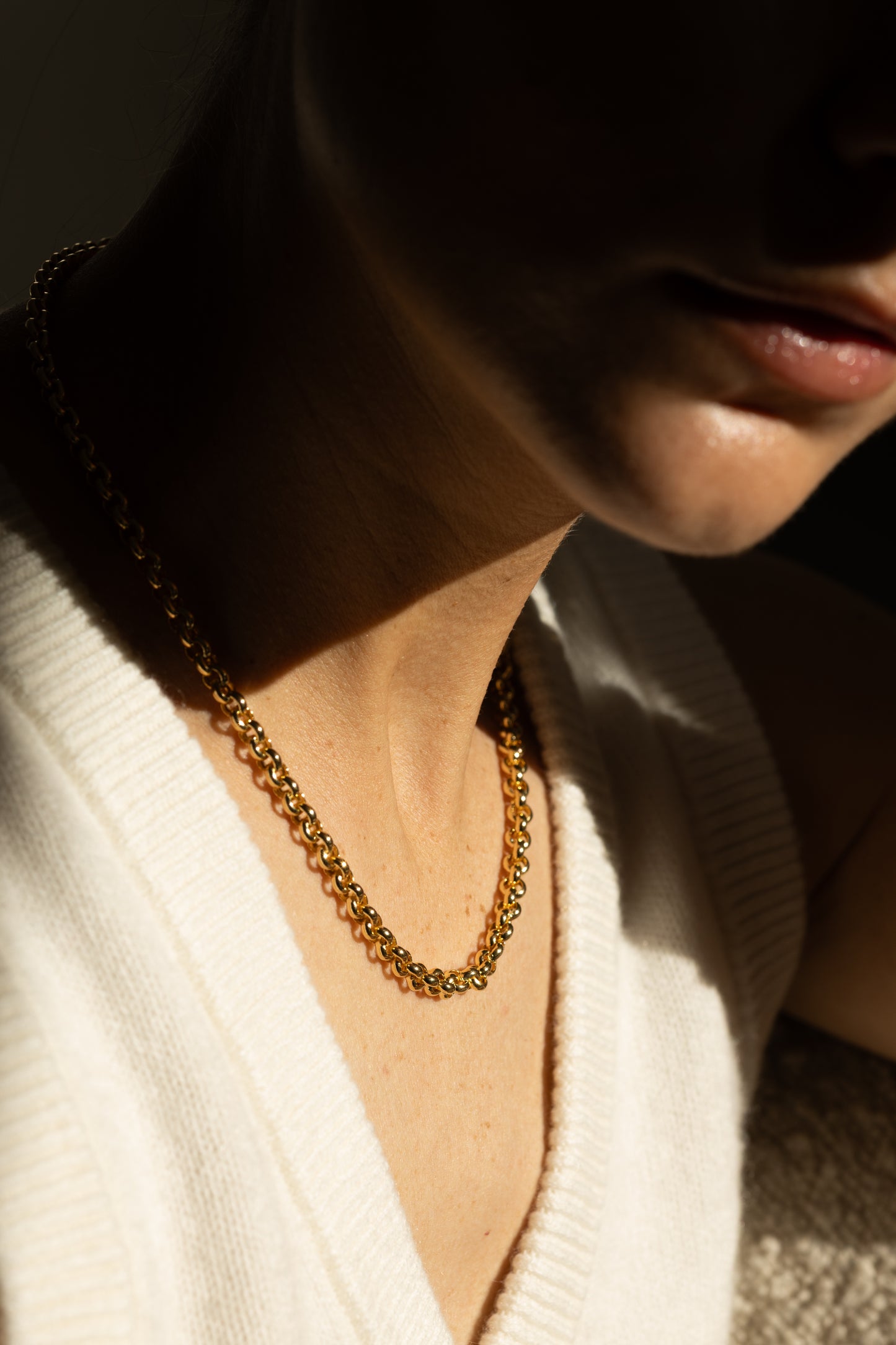 A close-up lifestyle shot of a woman wearing a statement gold chain necklace, paired with a white ribbed knit top under dramatic, high-contrast sunlight.