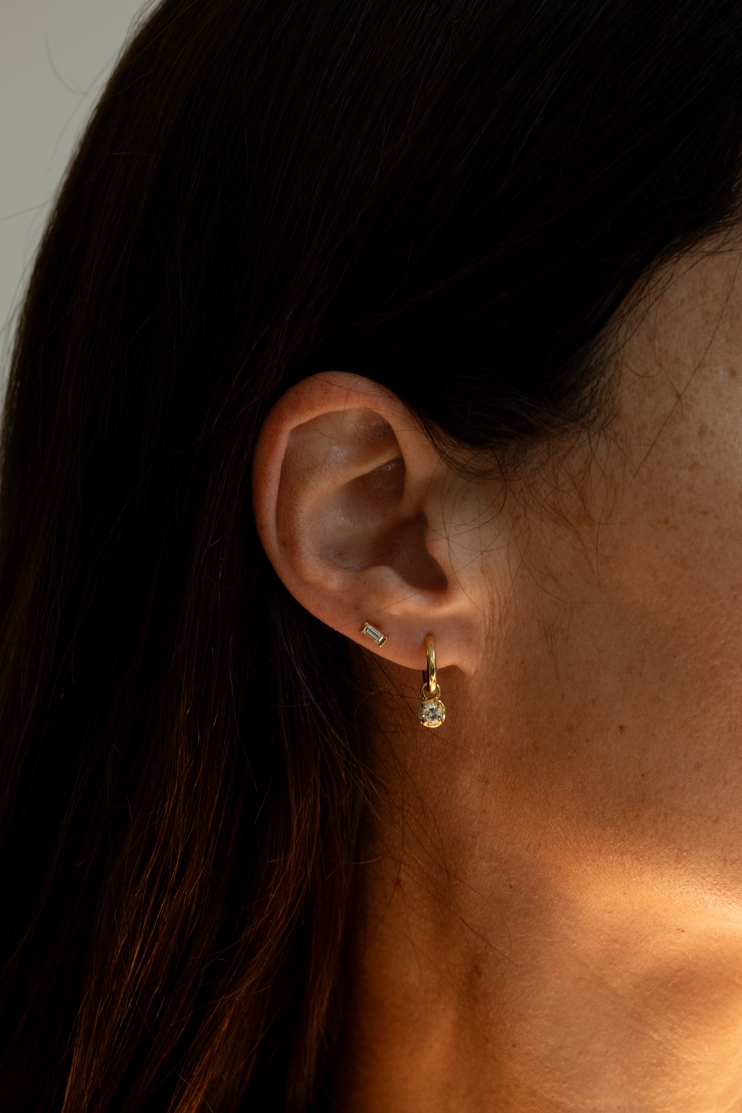 Close-up of a woman's ear wearing a small gold hoop earring with a tiny diamond and a small bugette diamond stud, against dark hair and warm light.