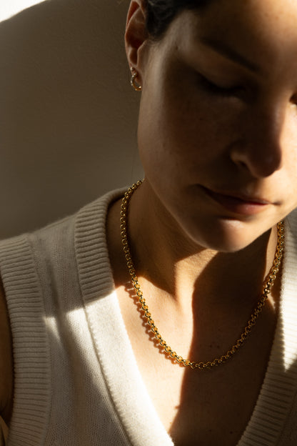 A close-up lifestyle shot of a woman wearing a statement gold chain necklace and a small gold hoop earring, dressed in a cream ribbed knit sweater under dramatic, warm side lighting.