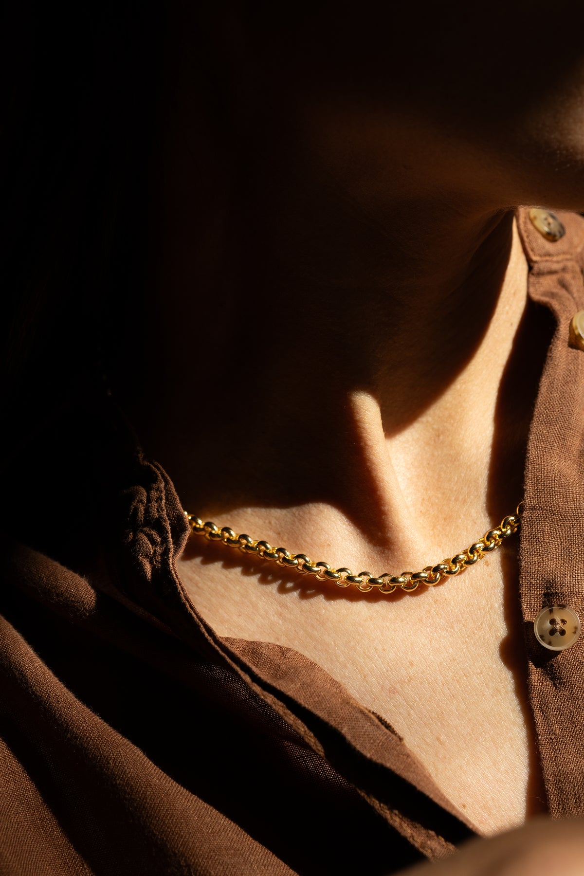 A close-up of a person wearing a statement gold chain necklace over a brown button-up shirt, captured in dramatic, high-contrast warm lighting.