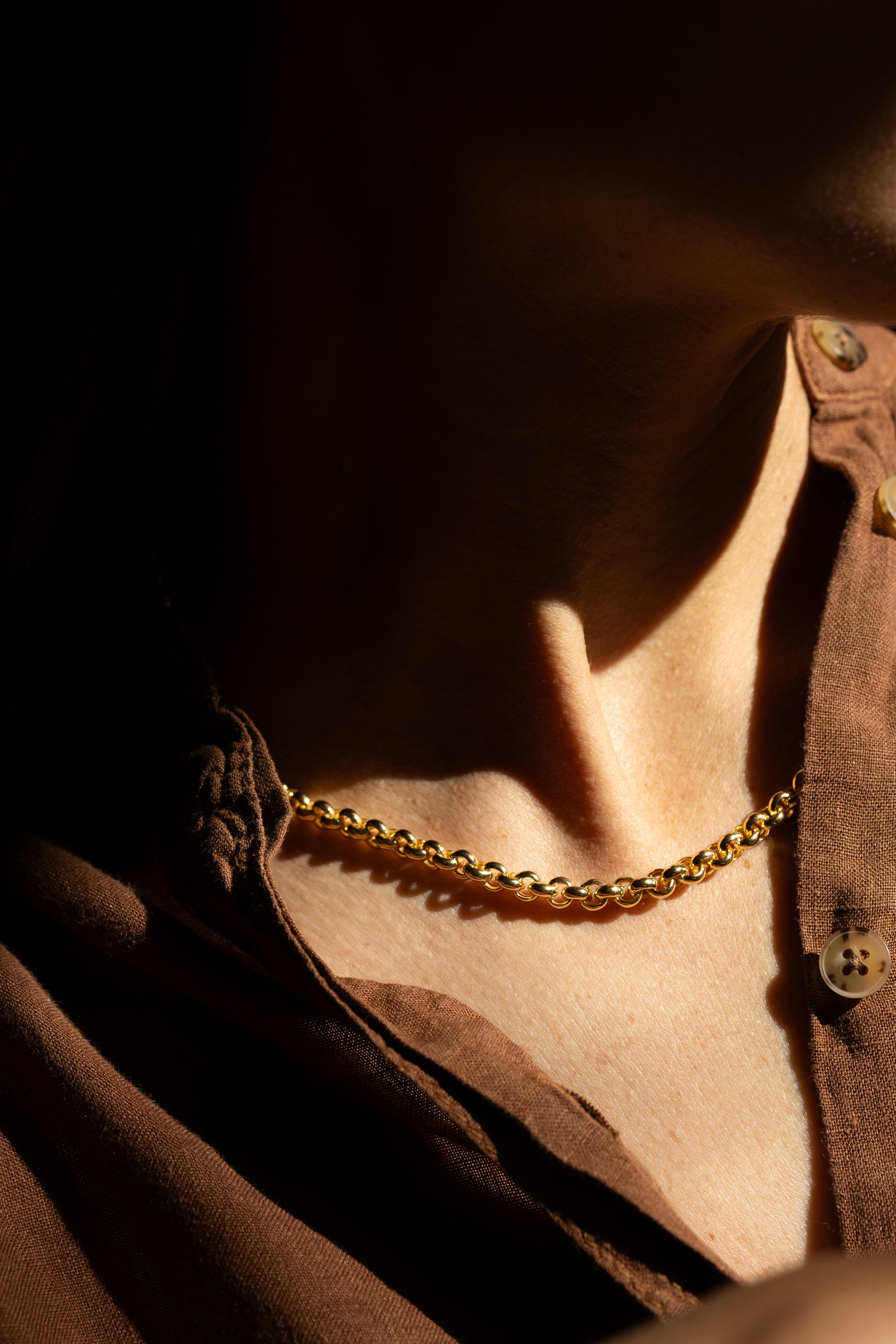 A close-up of a person wearing a statement gold chain necklace over a brown button-up shirt, captured in dramatic, high-contrast warm lighting.