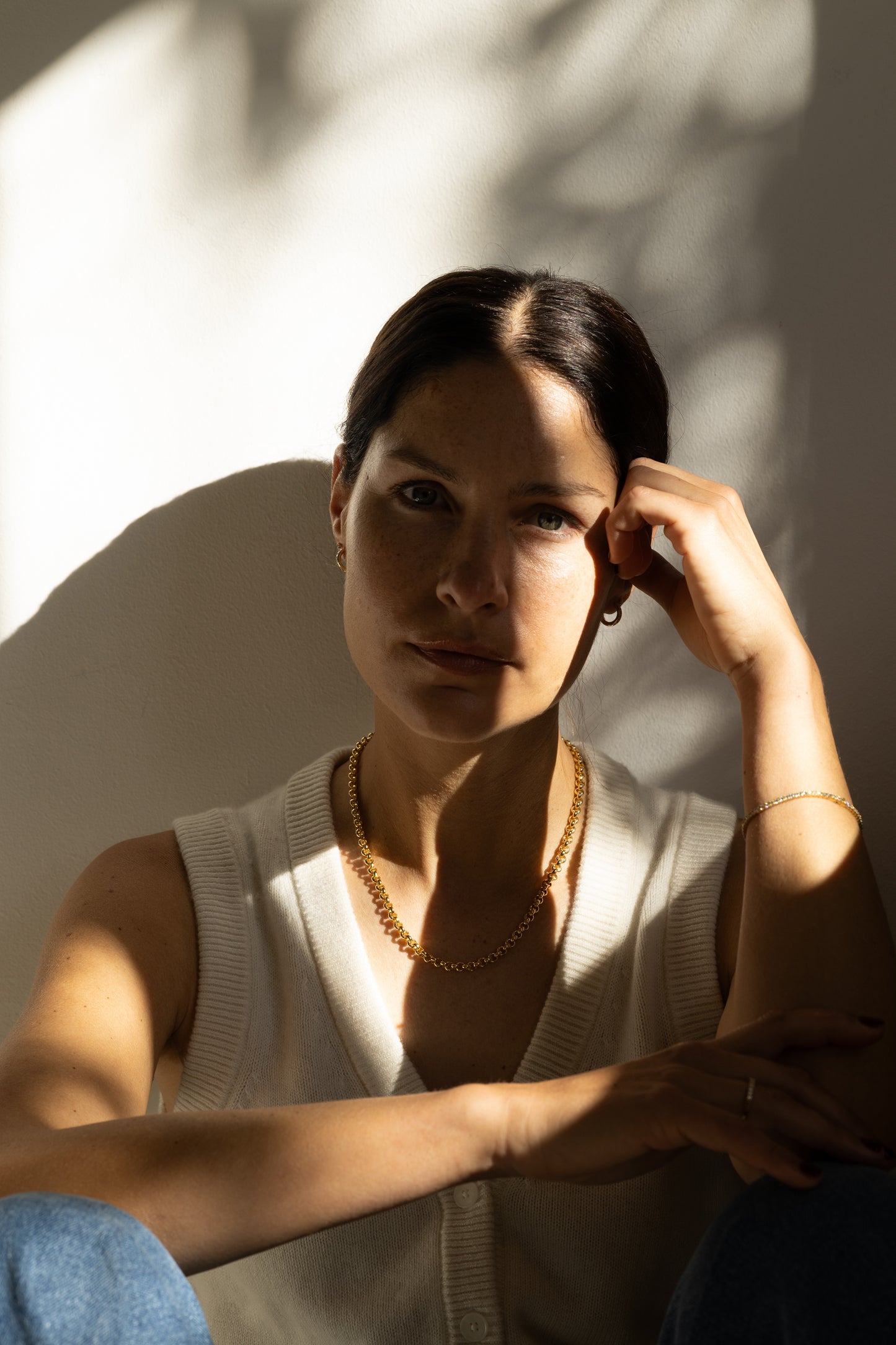 A woman wearing a gold chain necklace and diamond tennis bracelet, sitting in warm, high-contrast sunlight against a wall with leaf shadows.