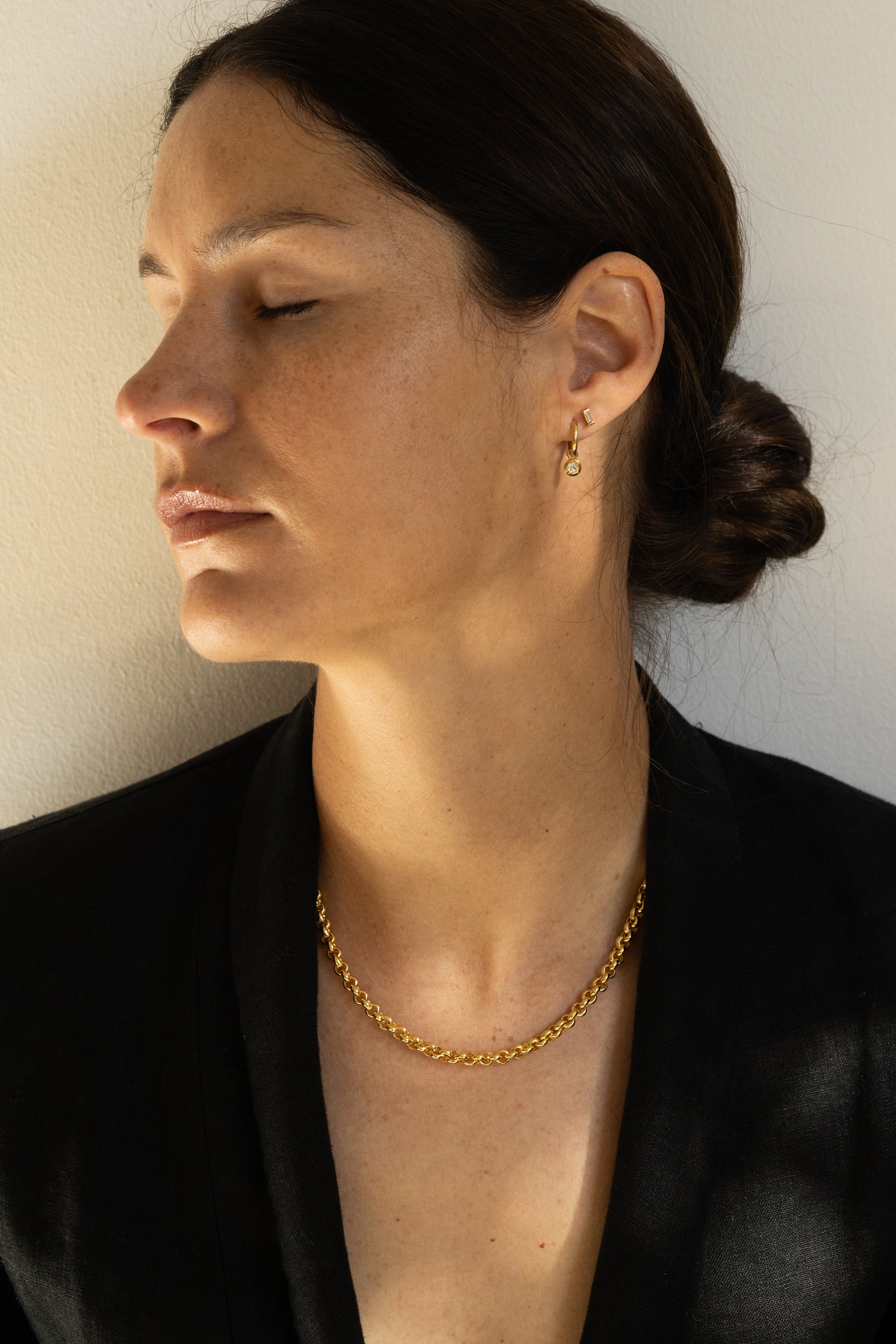 A woman in profile wearing a statement gold chain necklace and a small gold hoop earring, dressed in a black top against a plain light beige wall.
