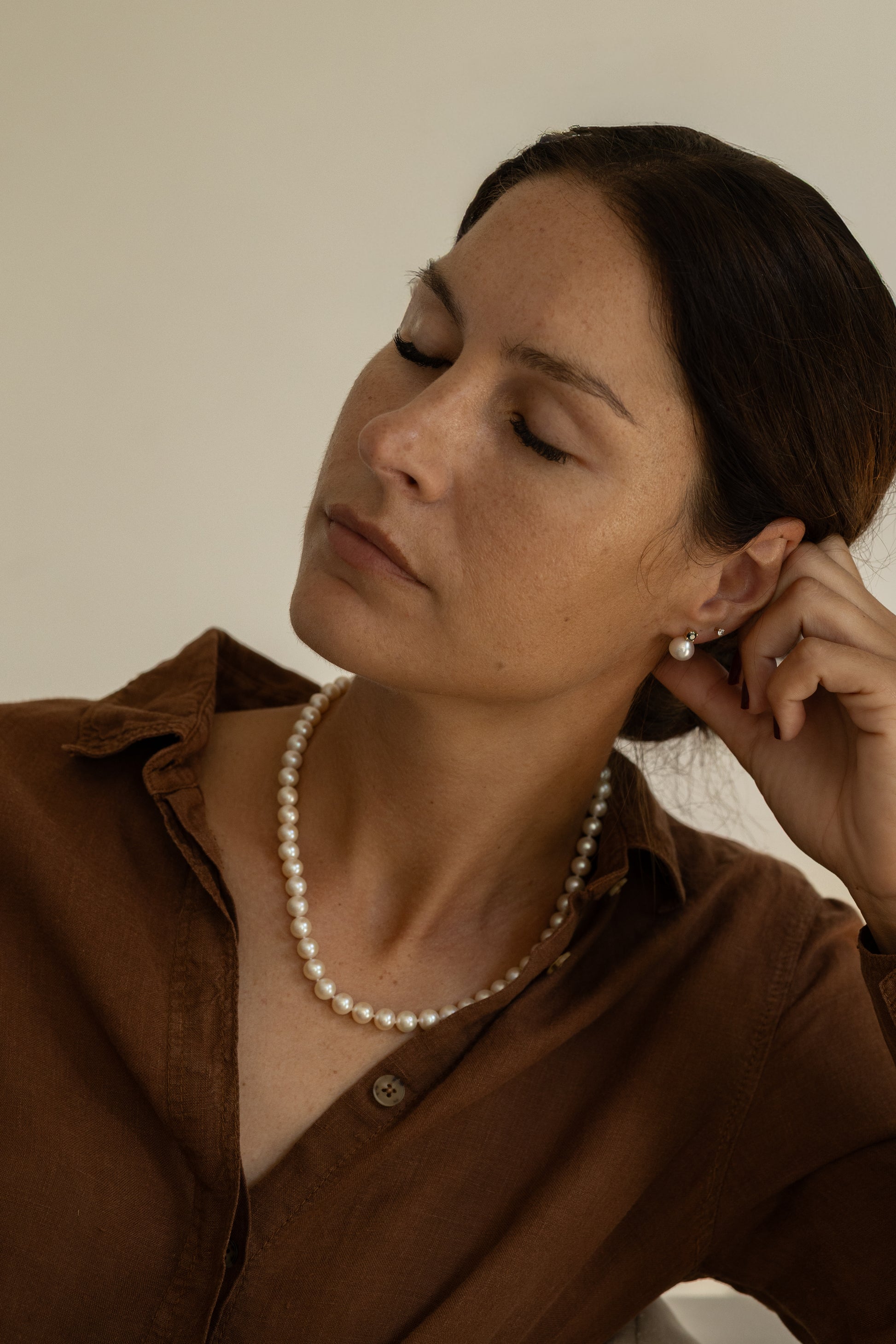 Woman wearing a pearl necklace and brown shirt against a neutral background