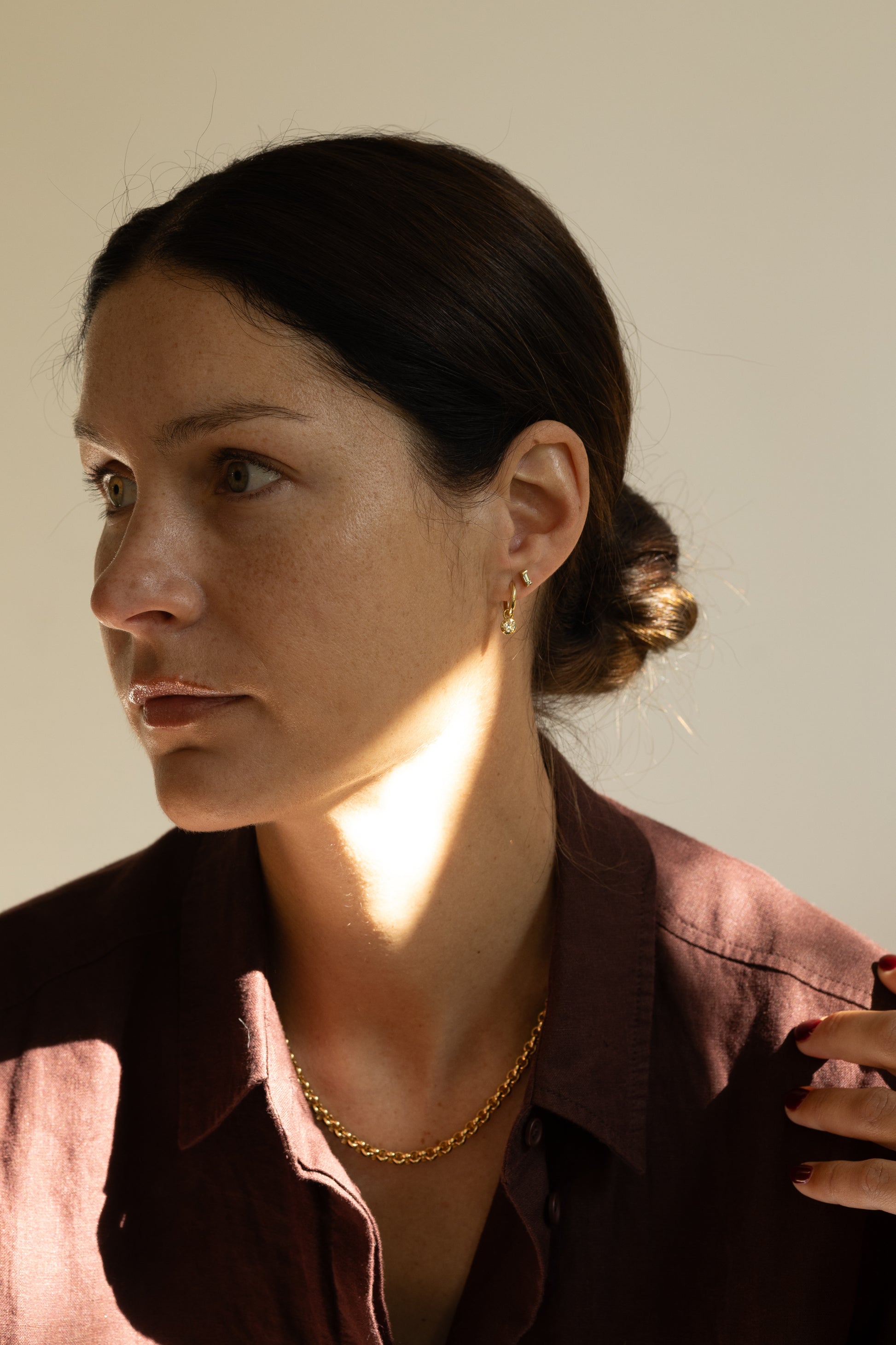 A woman with dark hair in a low bun wearing a gold chain necklace and a small gold hoop earring, wearing a dark reddish-brown collared shirt against a plain beige background.
