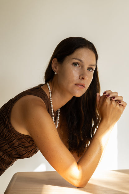 Woman with long dark hair and a pearl necklace sitting at a table with a warm light source.