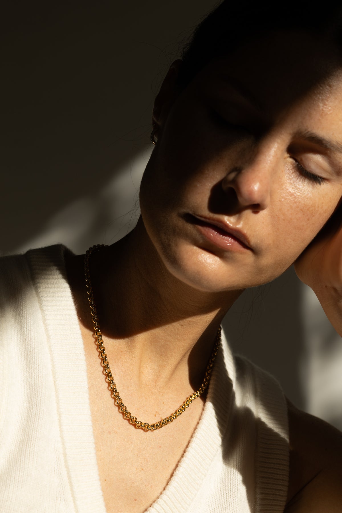 A close-up lifestyle shot of a woman with her eyes closed, wearing a statement gold chain necklace and a white ribbed knit top, set against a dark background with dramatic, high-contrast sunlight and shadows.
